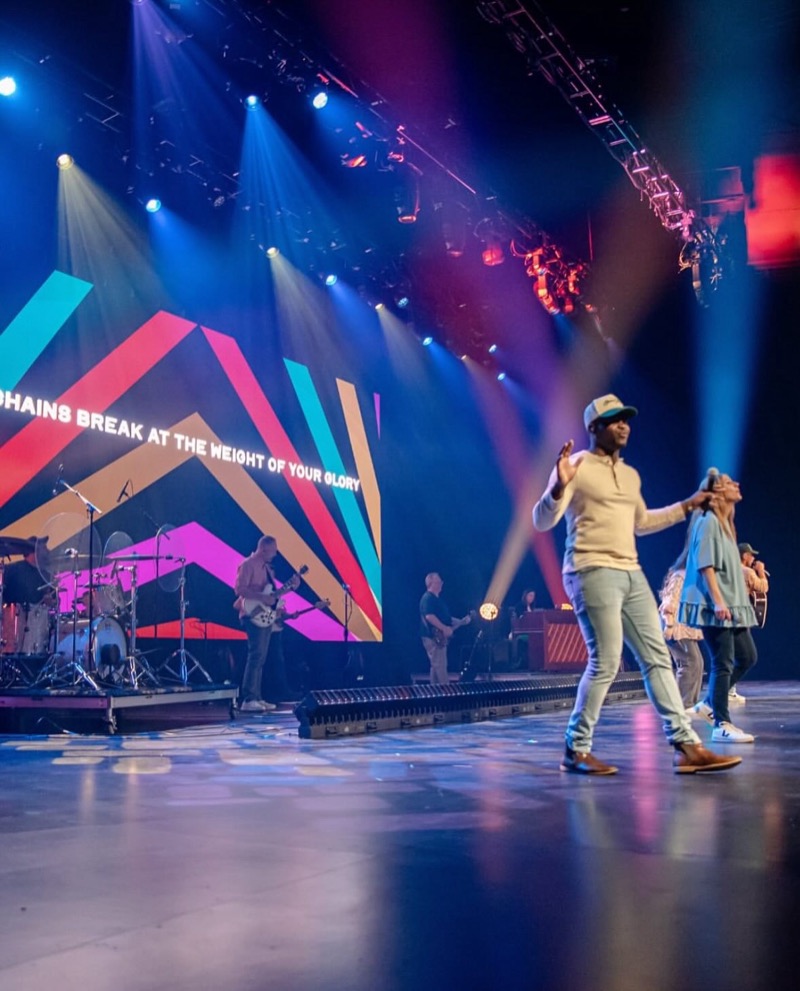 Vocalist crossing a reflective stage as blue beams and truss fixtures fill the air above.